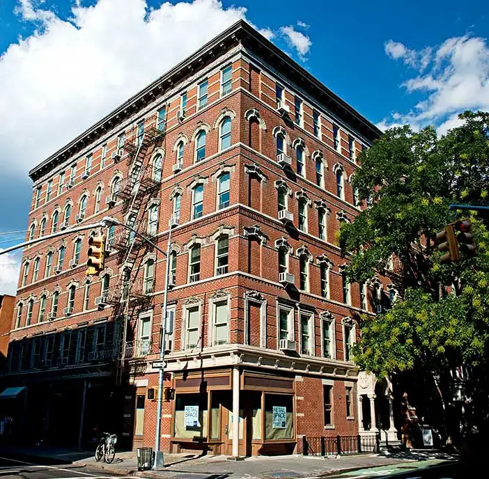 WindowFix - A five-story red brick apartment building with fire escapes on the corner of a city street under a partly cloudy sky.