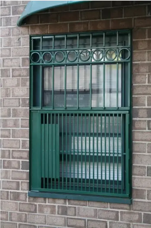 WindowFix - A brick wall with a window secured by ornate green metal bars and partially covered by vertical slats on the lower half.