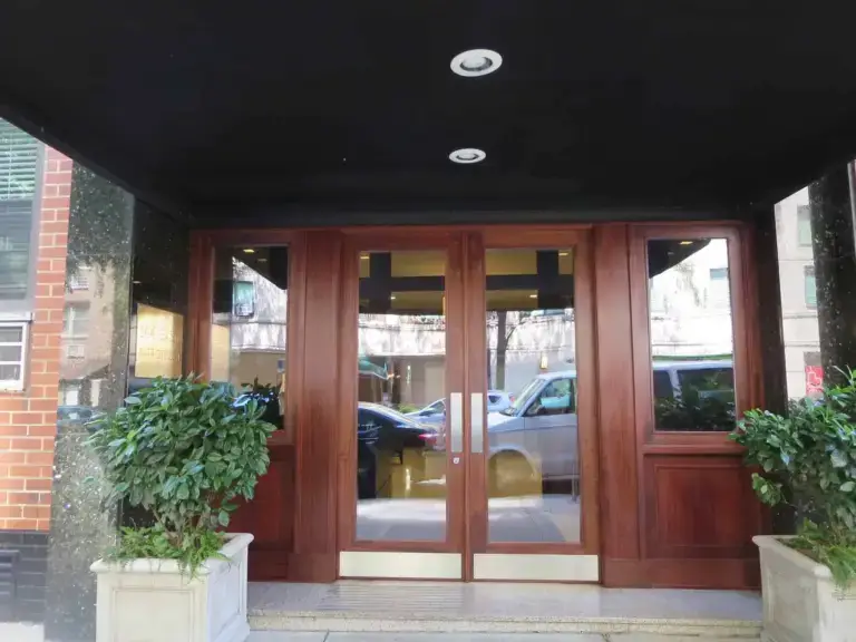 WindowFix - A double wooden door entrance with glass panels, flanked by large potted plants, leads into a building; cars are visible through the glass.