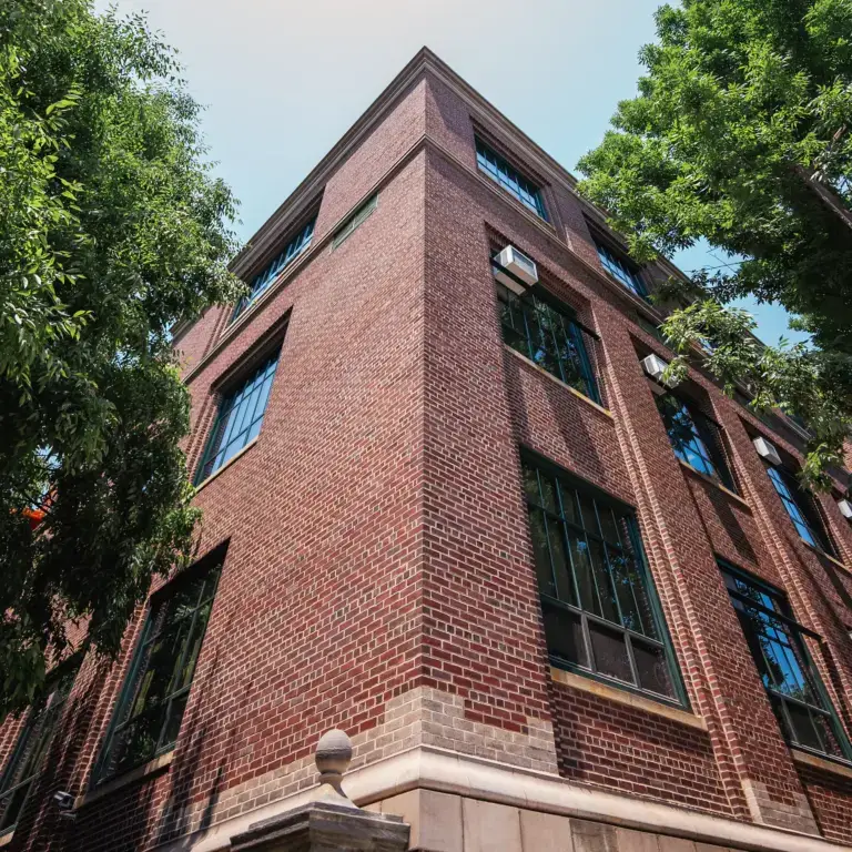 WindowFix - A three-story red brick building with large windows, viewed from a low angle, where newly install glass panels glisten, surrounded by green trees under a clear sky.