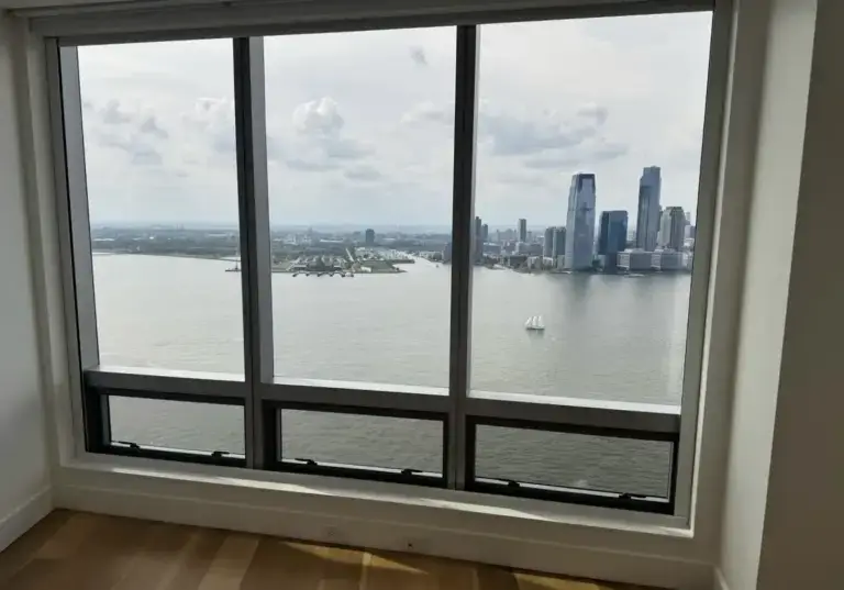 WindowFix - A large window offers a view of a river, distant city skyline with tall buildings, and a sailboat on the water under a partly cloudy sky.