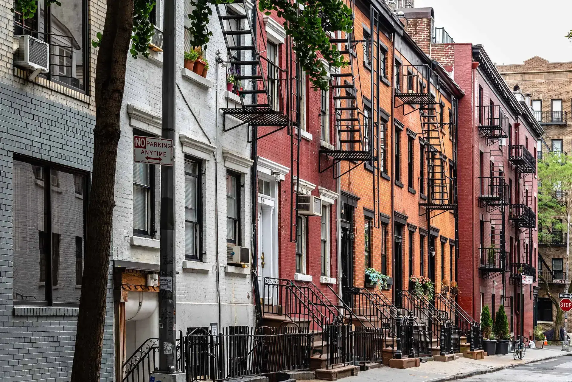 WindowFix - A row of brick and painted New York apartment buildings with fire escapes and stoops lines a quiet city street with a no parking sign and trees.