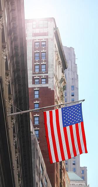 WindowFix - An American flag hangs from a building in a city, with sunlight and tall buildings visible in the background.